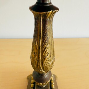 Pu&ograve; includere: Un candelabro decorativo con una parte superiore e inferiore marrone scuro e un centro dorato con un motivo a foglie. La base &egrave; quadrata con un bordo perlato. Il candelabro &egrave; in metallo e poggia su una superficie di legno chiaro.