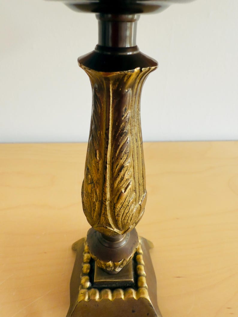 Pu&ograve; includere: Un candelabro decorativo con una parte superiore e inferiore marrone scuro e un centro dorato con un motivo a foglie. La base &egrave; quadrata con un bordo perlato. Il candelabro &egrave; in metallo e poggia su una superficie di legno chiaro.