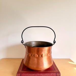 Può includere: Un calderone di rame con un manico nero, appoggiato su un libro rosso. Il calderone ha una forma arrotondata con una serie di cerchi decorativi. L'interno del calderone ha un aspetto più scuro e invecchiato.