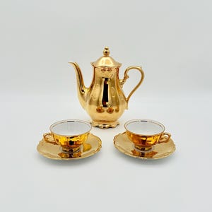 May include: A gold-colored tea set with a teapot and two teacups with saucers. The teapot has a curved spout and a lid with a decorative finial. The teacups and saucers have a scalloped edge and a white interior.