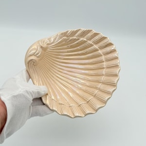 Può includere: Un piatto decorativo a forma di conchiglia, di colore pesca chiaro con riflessi iridescenti. Il piatto presenta una texture dettagliata e scanalata. È tenuto da una mano guantata su sfondo bianco.