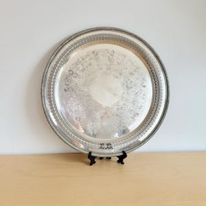 Puede incluir: Una bandeja redonda plateada con un diseño floral ornamentado en el centro. La bandeja tiene un borde decorativo y un borde en forma de cuerda. Se muestra sobre un soporte negro contra una pared blanca y una superficie de madera.