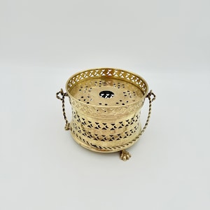 May include: A gold-coloured metal tea warmer with a pierced design. The tea warmer has a round shape with a decorative rim, two handles, and small feet. The design includes cut-out patterns and a braided rope detail.