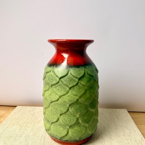 May include: A ceramic vase with a textured, scale-like pattern in shades of green. The top rim is a vibrant red, transitioning to green with a black band. The base is also red. The vase is displayed on a neutral-coloured surface.