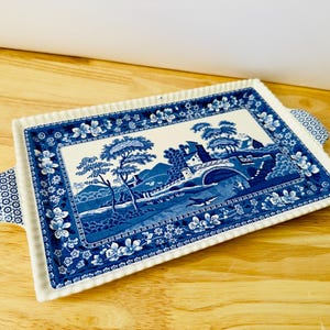 Pode incluir: Bandeja retangular de cerâmica com um design de paisagem azul e branco. A bandeja apresenta uma ilustração detalhada de uma ponte, árvores e edifícios. A borda é decorada com padrões florais e tem duas alças.