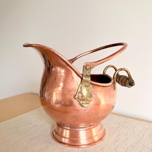 Puede incluir: Un recipiente de carbón de cobre con un pico curvo y un asa de latón. El recipiente tiene un cuerpo redondo y se asienta sobre una base de pedestal. El asa es de latón y madera. Probablemente es antiguo.