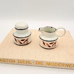 May include: A cream-colored ceramic sugar bowl with a lid and a small creamer. Both pieces have a speckled finish, dark blue trim, and a brown leaf design. The set is displayed on a light-colored surface.