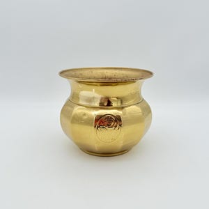 Puede incluir: Una pequeña olla de latón de estilo antiguo con un borde ancho y acampanado y un cuerpo redondeado. La olla tiene un emblema decorativo en la parte delantera y un acabado dorado pulido. La olla probablemente se utiliza con fines decorativos.