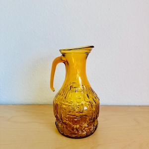 Op de afbeelding: Amberkleurige glazen kan met een gestructureerd, reliëf ontwerp. De kan heeft een handvat en een tuit om uit te schenken. Het glas heeft een warme, gouden tint en het ontwerp lijkt een natuurlijk thema te hebben. De kan is waarschijnlijk vintage.