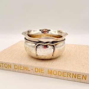 May include: A small, silver-coloured bowl with a hammered texture and a gold-coloured interior. The bowl is encircled by a gold-coloured rope design. The bowl sits on a book with gold lettering that reads "STON DIEHL - DIE MODERNEN".