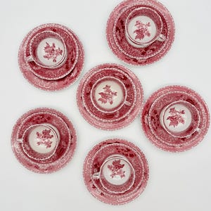 Puede incluir: Cinco juegos de tazas de té y platillos de cerámica dispuestos en un patrón circular. Cada pieza presenta un estampado floral de color rojo oscuro sobre un fondo blanco. Las tazas tienen platillos y platos a juego con motivos botánicos.