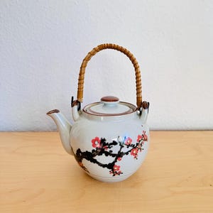 May include: A white ceramic teapot with a brown lid and a woven handle. The teapot is decorated with a floral design featuring red blossoms and black branches. The teapot is sitting on a wooden surface.