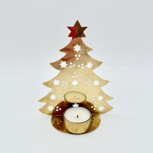 May include: A gold-colored metal tea light candle holder shaped like a Christmas tree. The tree features cut-out star and dot patterns throughout its surface and is topped with a star. A small, round, white tea light candle sits in the base.