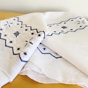 以下が含まれることがあります： 白のリネン生地に、青色の刺繍で施された複雑な花柄と幾何学模様。刺繍は生地の端に沿って装飾的なボーダーを形成し、繊細でエレガントなデザインを作り出しています。生地は折りたたまれ、詳細な職人技が示されています。