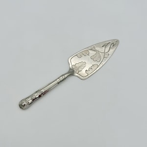 Può includere: Un server per torte d'argento con un manico ornato e una lama decorata con motivi a foglie. Il server è realizzato in metallo lucido ed è progettato per servire torte o crostate. Il design generale è elegante e vintage.