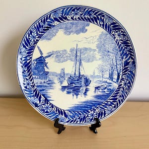 Könnte beinhalten: Runder, blau-weißer Zierteller mit einer Landschaft mit Windmühle und Booten auf dem Wasser. Der Teller hat einen detaillierten Rand mit Blumen- und Blättermustern. Der Teller wird auf einem schwarzen Ständer präsentiert. Durchmesser ca. 25 cm.
