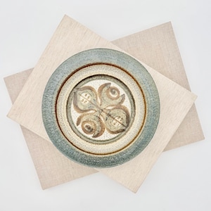 May include: A round ceramic bowl with a speckled cream-colored center featuring a brown, four-petaled floral motif. The bowl has a wide, textured rim in a muted blue-green glaze, creating a distinct contrast against the lighter interior base.