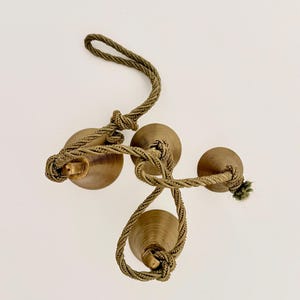 May include: Three bronze-colored decorative bells are connected by a twisted, olive-green rope. The bells have a rounded shape and are arranged in a cluster. The rope is knotted in several places, creating a decorative element.