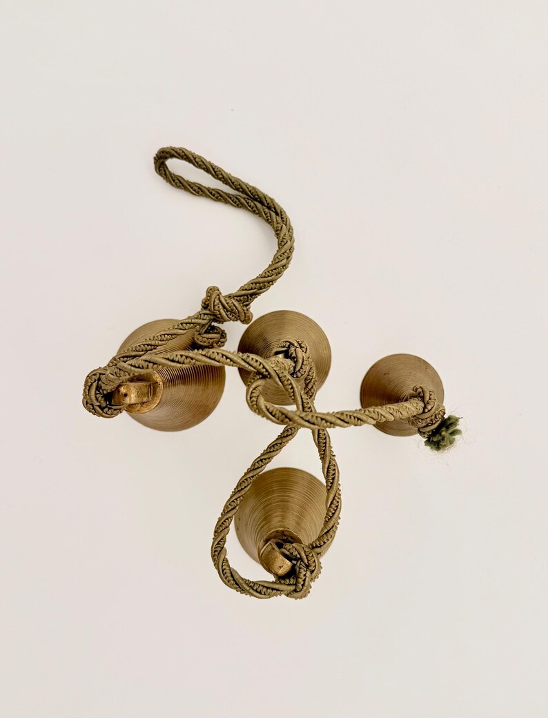 May include: Three bronze-colored decorative bells are connected by a twisted, olive-green rope. The bells have a rounded shape and are arranged in a cluster. The rope is knotted in several places, creating a decorative element.