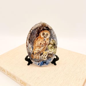 Könnte beinhalten: Ein ovales Kunstwerk aus Kunstharz mit einer detaillierten Eule. Die Eule ist in Braun- und Beigetönen dargestellt und sitzt auf einem Ast in einer geodenartigen Umgebung. Das Kunstwerk wird auf einem schwarzen Ständer präsentiert.