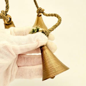 May include: A set of three brass bells with a textured, ribbed design. The bells are suspended from a twisted rope. The bells are held by a person wearing a white glove.