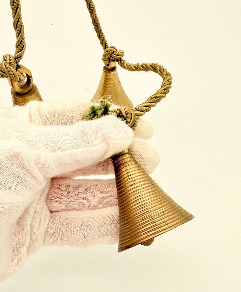 May include: A set of three brass bells with a textured, ribbed design. The bells are suspended from a twisted rope. The bells are held by a person wearing a white glove.