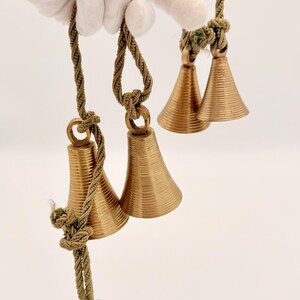 May include: Close-up of three sets of small, gold-colored bells with a ridged design. Each bell is suspended by a twisted, olive-green rope. The bells are held by a gloved hand against a white background.