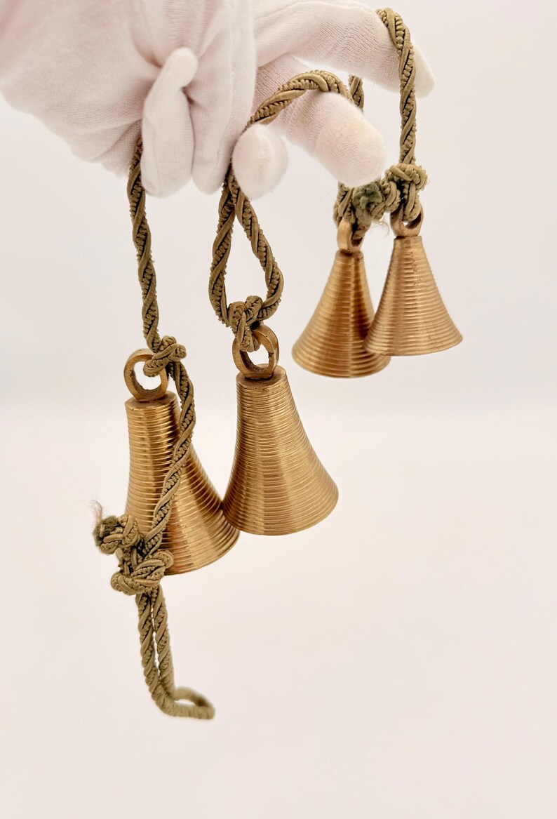 May include: Close-up of three sets of small, gold-colored bells with a ridged design. Each bell is suspended by a twisted, olive-green rope. The bells are held by a gloved hand against a white background.