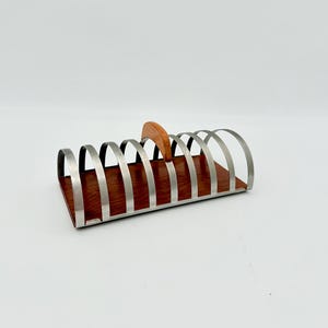 May include: A rectangular toast rack featuring a dark brown wooden base and a series of arched, brushed metal dividers. A small, curved wooden handle is positioned at the center of the top, providing a grip for the metal structure.