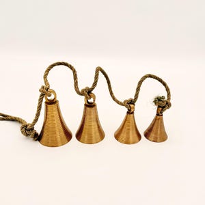 May include: Four bronze-colored bells of varying sizes are strung together on a twisted rope. The bells have a conical shape with horizontal ridges. The rope is knotted between each bell and at the end, creating a decorative hanging ornament.