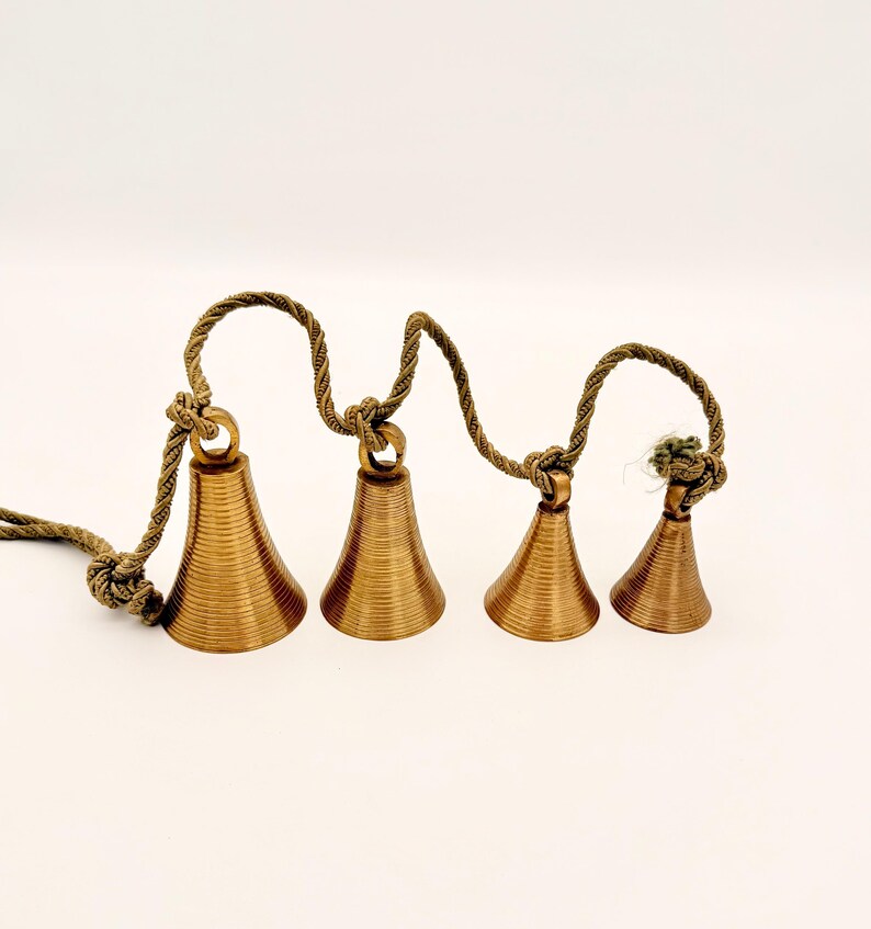 May include: Four bronze-colored bells of varying sizes are strung together on a twisted rope. The bells have a conical shape with horizontal ridges. The rope is knotted between each bell and at the end, creating a decorative hanging ornament.