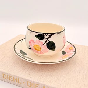 Op de afbeelding: Crèmekleurige theekop en schotel set met roze bloemmotieven en groene bladeren. De kop en schotel hebben een zwarte rand. De set staat op een lichtgekleurd boek met gouden letters.