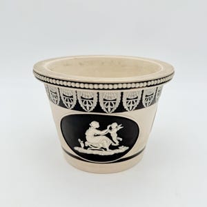 May include: A cream-colored ceramic planter featuring a wide, beaded rim and a black oval medallion on the front. The medallion displays a white relief scene of a seated figure interacting with a winged cherub, accented by a decorative border.