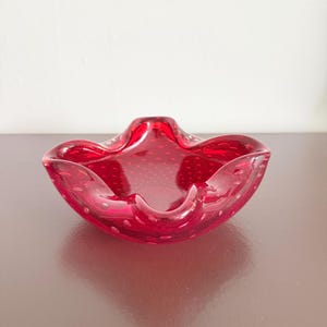 以下が含まれることがあります： 厚いガラス壁の中に多数の小さな気泡が浮かぶ、独特のブリカンテ技法を用いた鮮やかな赤色のガラスボウル。ボウルには、複数の曲線を描く幅広で波打ったスカラップ状の縁があり、中央は深くなっています。