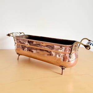 Peut inclure: Une jardinière rectangulaire en cuivre avec une finition polie, dotée de quatre petits pieds en métal et de deux poignées en laiton sur les côtés. Chaque poignée est ornée d'un insert en céramique blanche avec un motif floral bleu.