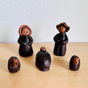 Könnte beinhalten: Ein Set aus fünf dunkelbraunen Tonfiguren, bestehend aus zwei Erwachsenenfiguren, einer zentralen Säuglingsfigur und zwei kleinen Tierfiguren. Jedes Stück hat eine glatte, polierte Oberfläche mit einfachen, handgeschnitzten Gesichtern.