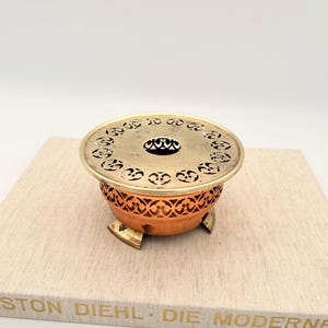 Könnte beinhalten: Ein Kupfer- und Messing-Räuchergefäß mit dekorativem durchbrochenem Design. Der Brenner hat eine runde Oberseite mit einer zentralen Öffnung und steht auf drei kleinen Füßen. Der Sockel ist aus Kupfer und die Oberseite aus Messing.