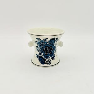 Könnte beinhalten: Ein cremefarbenes Keramikgefäß mit ausgestelltem Rand und Boden, das auf der Vorderseite mit einem dunkelblauen Blumenmuster bemalt ist. Das Stück hat zwei kleine, abgerundete Griffe an den Seiten und dünne blaue Linien.