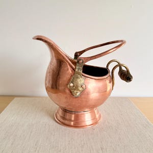 May include: A polished copper coal scuttle with a rounded body, a wide pouring spout, and a circular pedestal base. It features a brass swing handle attached with decorative plates and a smaller rear handle with a dark wooden grip for pouring.