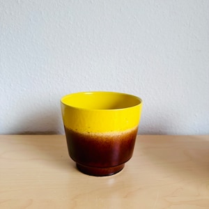 Può includere: Un vaso di ceramica con una parte superiore gialla che sfuma in una base marrone. Il vaso ha una forma cilindrica con un bordo leggermente svasato e una piccola base arrotondata. Il vaso è su una superficie di legno chiaro.