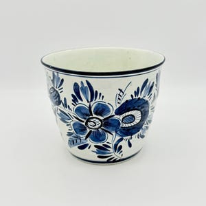 May include: A white ceramic planter with a blue floral design. The planter has a rounded shape with a black rim and is decorated with stylized blue flowers and leaves. The interior is a light green color.