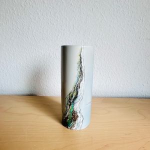 May include: A cylindrical ceramic vase with a light gray base color. The vase features a vertical abstract design with green, brown, and black accents. The design resembles a natural landscape. The vase is displayed on a light wood surface.