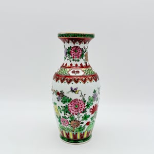 Pode incluir: Um vaso de cerâmica branco com a parte superior e inferior alargadas, adornado com desenhos florais e de borboletas coloridos. O vaso apresenta uma borda e base estampadas em vermelho e verde, com flores rosa, roxas e amarelas. Uma peça decorativa para casa.