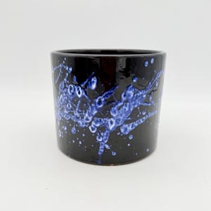 May include: A cylindrical ceramic planter with a glossy black finish featuring a vibrant blue drip glaze pattern that resembles splattered paint. The vessel has a smooth, uniform rim and a deep, dark base colour that contrasts with the bright blue.