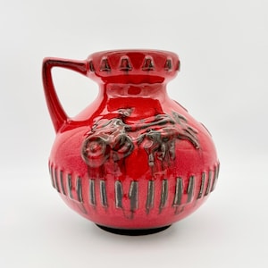 Puede incluir: Una jarra de cerámica de color rojo brillante con cuerpo redondeado y una sola asa. El recipiente presenta un patrón en relieve texturizado de color marrón oscuro de un carruaje tirado por caballos, con rayas verticales en la base.