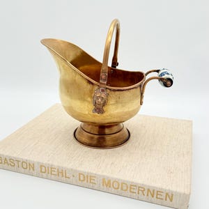 Peut inclure: Une ancienne huche à sucre en laiton avec une poignée et un visage décoratif à l'avant. La poignée latérale a un bouton en porcelaine bleue et blanche. La huche est posée sur un livre avec des lettres dorées.