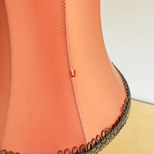 Pu&ograve; includere: Primo piano di un paralume color corallo con finiture decorative. Il paralume ha una consistenza liscia e una cucitura verticale. La finitura &egrave; di colore scuro con un motivo dettagliato. Il paralume &egrave; su una superficie chiara.