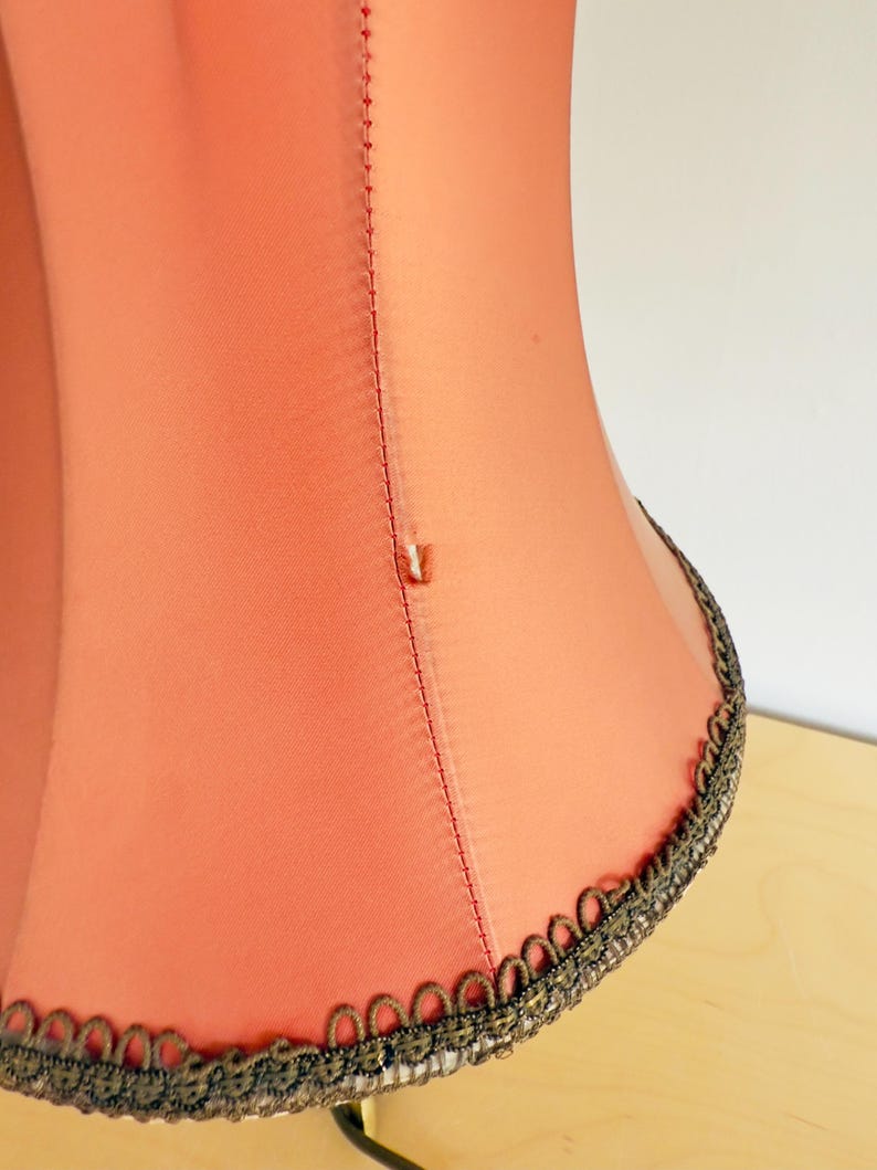 Pu&ograve; includere: Primo piano di un paralume color corallo con finiture decorative. Il paralume ha una consistenza liscia e una cucitura verticale. La finitura &egrave; di colore scuro con un motivo dettagliato. Il paralume &egrave; su una superficie chiara.