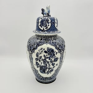 Puede incluir: Un jarrón de cerámica azul y blanco con tapa, con diseños florales y geométricos. El jarrón tiene un diseño detallado con una pequeña figura en la parte superior de la tapa. La estética general es tradicional y decorativa.