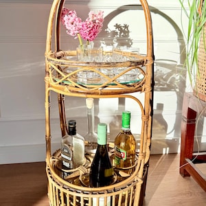 May include: A three-tiered, natural rattan bar cart with a handle. The top shelf holds clear glasses and pink flowers in a vase. The middle shelf has bottles, and the bottom shelf has bottles and a mirrored surface. The cart is on a wooden floor.